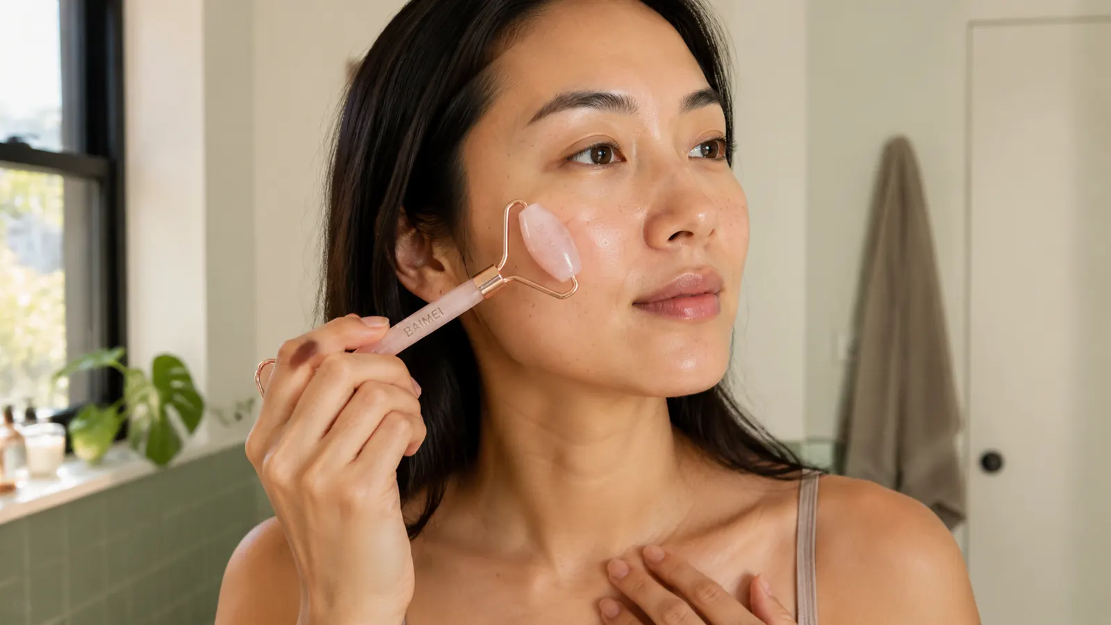 Hand holding the BAIMEI rose quartz roller against a woman's cheekbone, roller gliding upward toward the temple