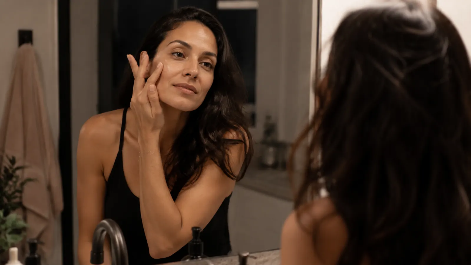 Woman applying a thin layer of clear serum to her cheek in a bathroom mirror at night