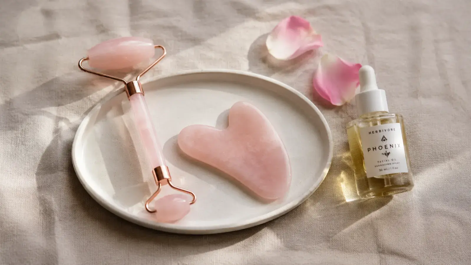 Rose quartz gua sha stone and roller set resting on a jade green tray next to a small bottle of facial oil