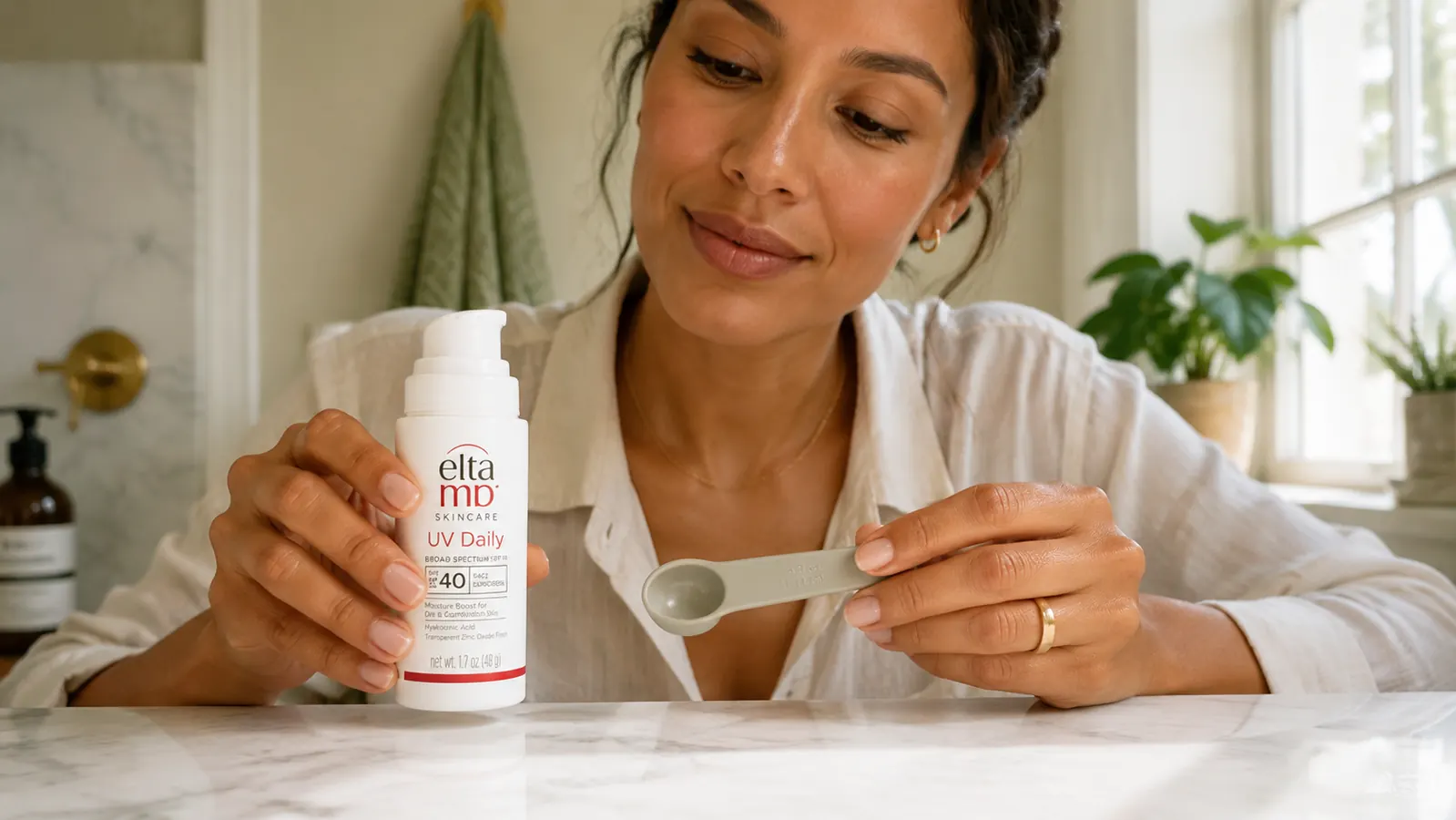 Hand holding a pump bottle of EltaMD UV Daily SPF 40 next to a small teaspoon measuring tool on a marble surface