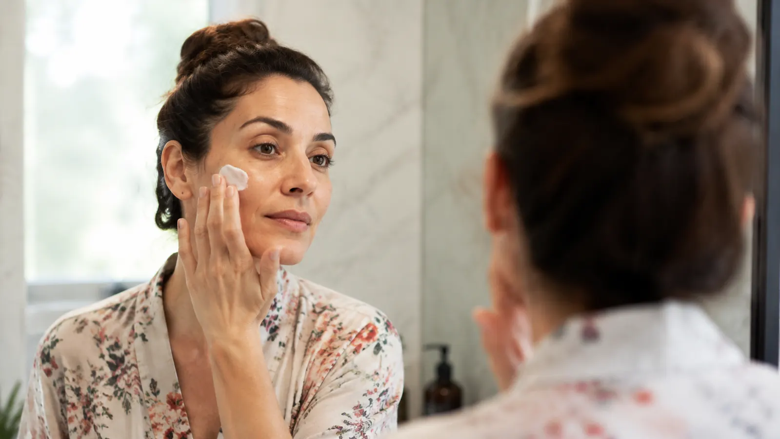Woman applying moisturizer to cheek in bathroom mirror, relaxed morning routine