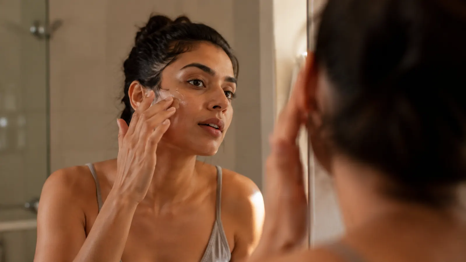 Woman with medium-light skin applying serum to her cheek in front of a bathroom mirror, morning light, relaxed expression, no heavy makeup