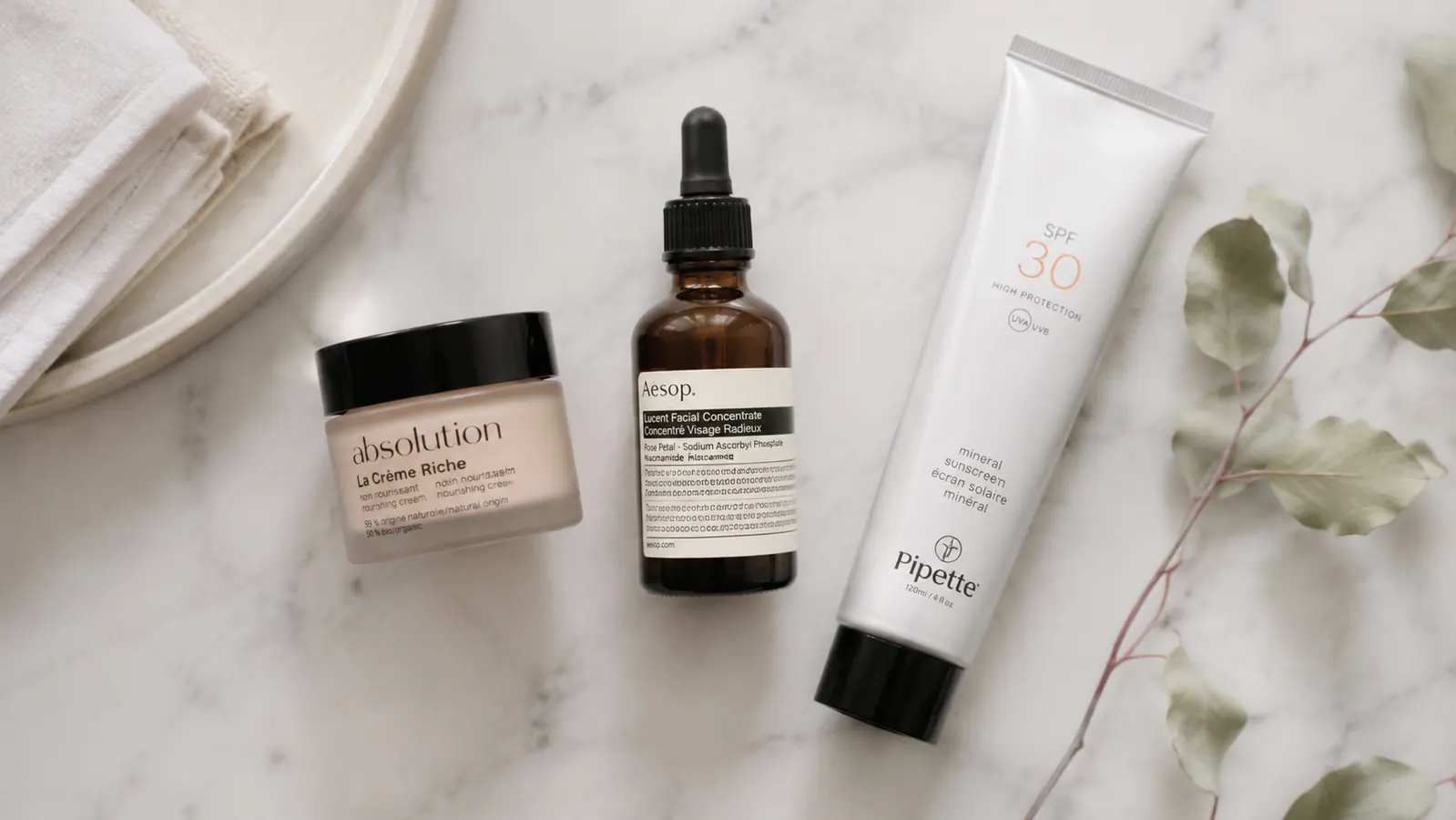 Skincare flatlay showing a vitamin C serum bottle, moisturizer, and SPF arranged on a marble surface with small green plant